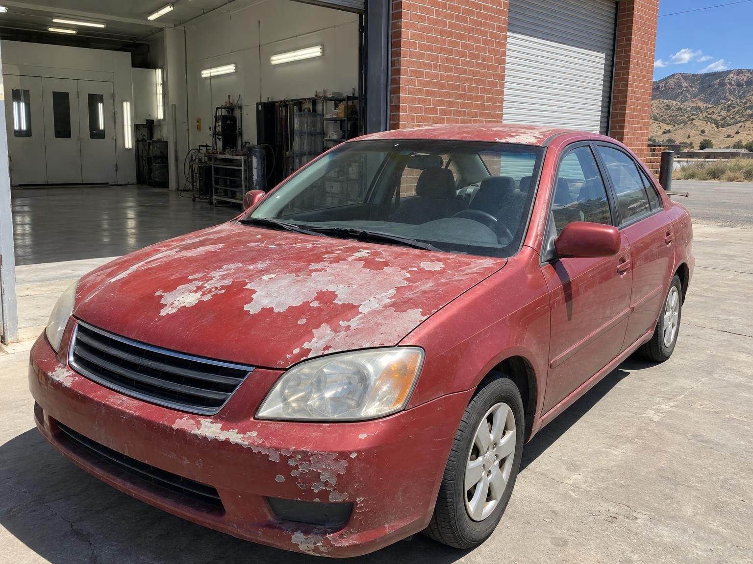 Red compact sedan with sun-peeled clear coat on the hood and bumper outside the open bay