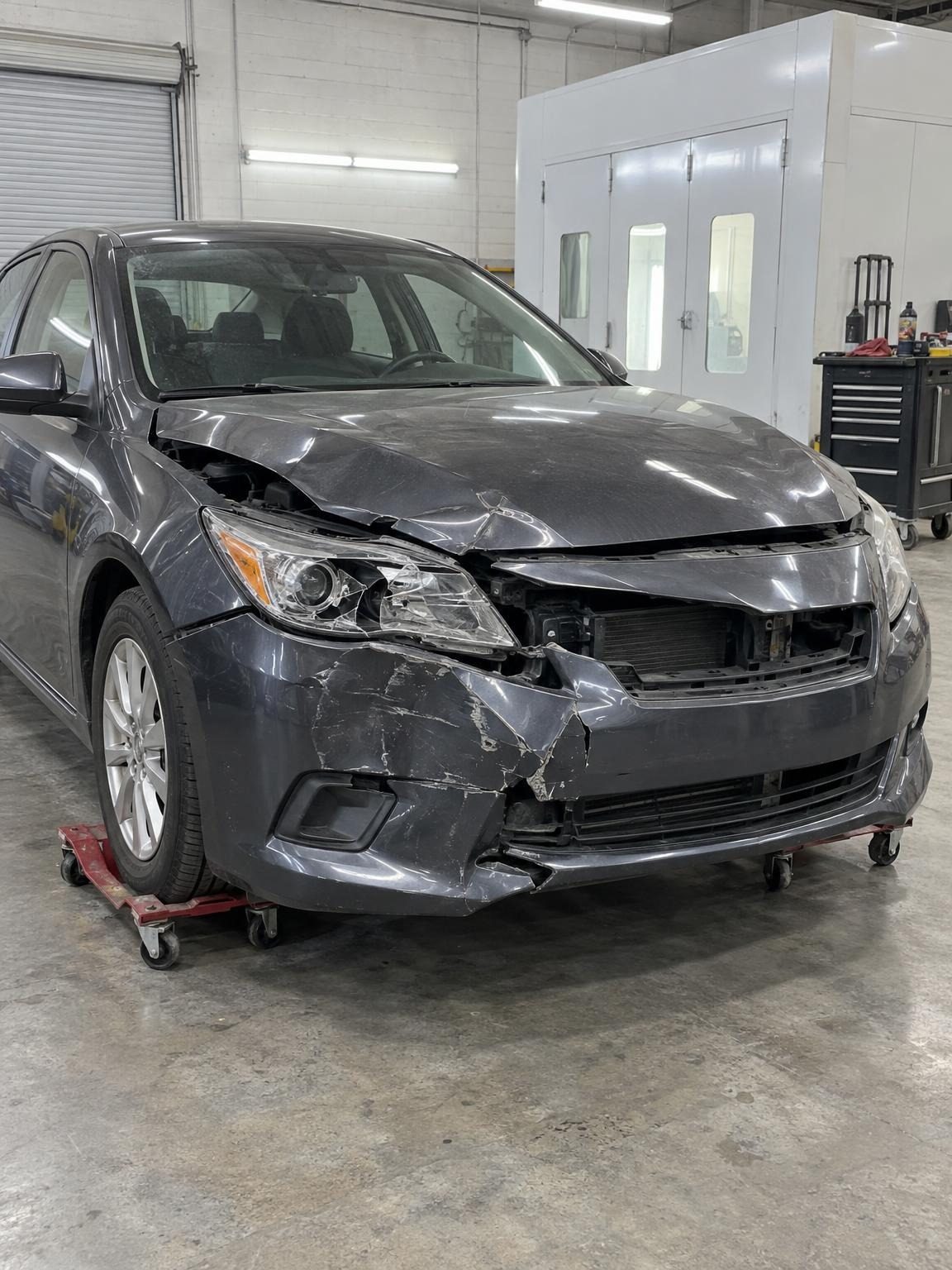 Dark grey compact sedan with front-end collision damage on wheel dollies in the bay